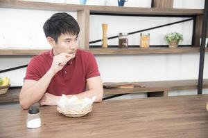 un joven asiático con una camiseta roja casual disfruta desayunando, comiendo papas fritas. joven cocinando comida en la cocina estilo loft foto