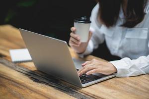 primer plano de la mano de una mujer de negocios que trabaja en una laptop y sostiene una taza de café caliente en una mesa de madera con una taza de café. el concepto de negocio trabaja desde casa en cualquier lugar. foto