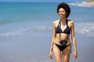 Joyful black woman with photo camera on wet coast