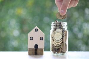 Model house on stack of coins money and woman hand putting a coin into the glass bottle on natural green background,Business investment and real estate concept photo