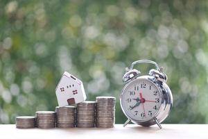 Model house on stack of coins money and alarm clock on natural green background, investment and business concept photo