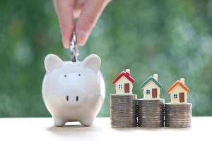 Woman hand putting a coin into piggy bank with model house on stack of coins money on nature green background,Save money for prepare in future and real estate concept photo