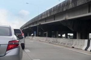 Rear side of silver car waiting in line for other cars. Cars on the concrete road with side crossing bridge. Under the blue sky with white car at days. photo