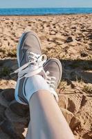 Legs in gray sneakers on beach sand. Summer vacation concept by the sea. Lifestyle travel. Pov view. Copy space. photo