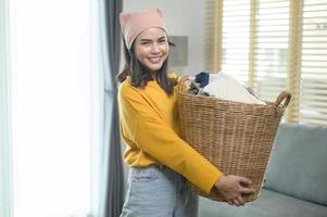 joven mujer feliz con pantalones amarillo sosteniendo una cesta llena de ropa en casa, concepto de lavandería foto