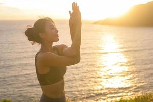 Young asian woman in sportswear doing yoga on the rock at seaside during Sunset, health and meditation concept photo