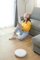 Young happy woman relaxing and using smartphone in living room while Robotic vacuum cleaner working photo