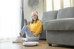 joven mujer feliz relajándose y usando un teléfono inteligente en la sala de estar mientras la aspiradora robótica funciona foto