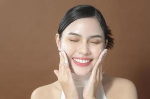 una mujer joven con cara hermosa sonriendo, lavándose la cara sobre fondo marrón, concepto de cuidado de la piel de belleza foto