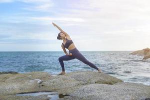 joven mujer asiática en ropa deportiva haciendo yoga en la roca en el concepto de playa, salud y meditación foto