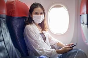 mujer joven con mascarilla está usando un teléfono inteligente a bordo, nuevo viaje normal después del concepto de pandemia covid-19 foto