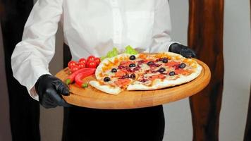 Delicious pizza on wooden tray in restaurant kitchen. Crop view of chef's hands in black gloves holding tasteful pizza with vegetables next to, spinning cutting board in pizzeria. Concept of food video