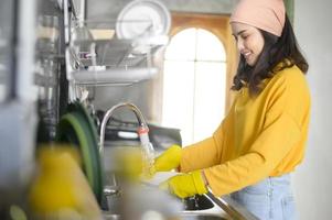 joven mujer feliz usando guantes amarillos lavando platos en la cocina en casa foto