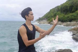 joven en ropa deportiva haciendo arte marcial, qigong, tai chi en la roca en la playa, concepto de salud y meditación foto