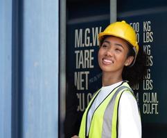 Smiling beautiful African American female foreman worker wears yellow safety hardhat watching cargo boxes loading at container yard, portrait. Industrial engineer woman works at international dockyard photo