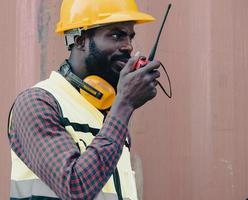 African American foreman holds walkie talkie for remote communication with teamwork. Industrial engineer wearing helmet and reflective vest working at container yard, import export shipping terminal. photo