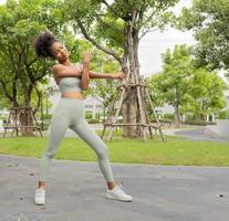 Young black woman stretching, exercising in nature park. Beautiful girl in sportswear warming by physical exercise for flexibility of muscles before jogging run in green garden. Healthy lifestyle girl photo