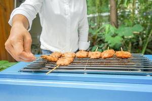 chica asiática de camiseta blanca sosteniendo y poniendo palitos de cerdo a la parrilla en una máquina de estufa de parrilla azul frente al fondo del jardín de árboles borrosos al aire libre. foto