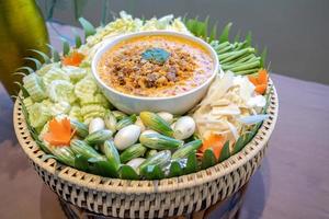 Traditional Thai chilli spicy sauce called Nam Prik in white bowl in the middle of vegetable cabbage cucumber carrot eggplant lentils ginger. photo