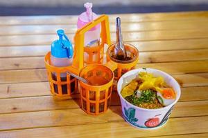 Asian noodle style in the paper bowl with flavoring beside on the wood table in the night. photo