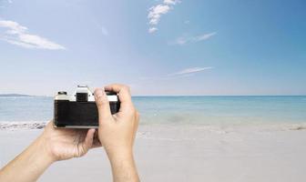 Man holding film camera ready to take photo over sea beach with blue sky background