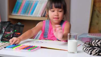 süßes kleines Mädchen, das Milch trinkt, während es eine Pause vom Zeichenunterricht macht. Asiatisches kleines Mädchen, das online lernt und Homeschooling genießt. Kind macht Hausaufgaben video