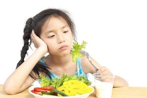 encantadora chica asiática que muestra una expresión aburrida con verduras frescas y coloridas y un vaso de leche aislado sobre fondo blanco foto