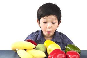 Chico sano asiático mostrando expresión feliz con variedad de frutas y verduras de colores sobre fondo blanco. foto