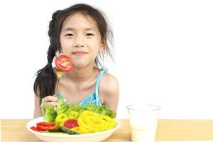 Encantadora chica asiática que muestra disfrutar de la expresión con verduras frescas y coloridas y un vaso de leche aislado sobre fondo blanco. foto