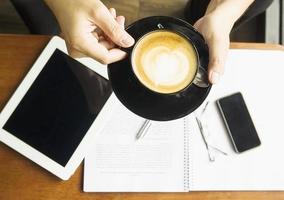 Man holding cup of coffee while working with his report in coffee shop. Photo is focused at coffee cup.