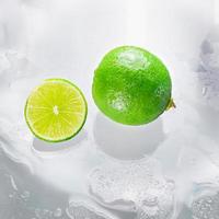 la lima verde está en blanco con la rodaja de lima cortada mostrando el interior de la pulpa de limón húmeda sobre una superficie de vidrio transparente, reflejando las sombras de la lima y el agua húmeda, dándole su frescura.
