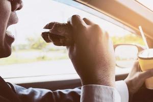 Man driving car while holding a cup of cold coffee and eating hamburger photo