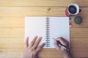 Top view of man writing note book on wooden table with coffee cup and small cactus pot photo