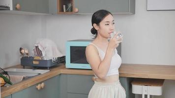mujer asiática en ropa deportiva bebiendo agua después de hacer ejercicio en casa una joven atlética bebe agua mineral natural limpia en una taza después de un entrenamiento para mantenerla saludable en la cocina de su casa. video