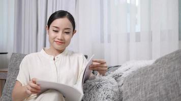 bella donna sorridente che legge un libro e si siede sul divano in soggiorno divertendosi a casa. video