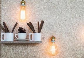 Cup of on cinnamon and pine cone on wooden shelves over wood pulp board background with warm light bulb photo