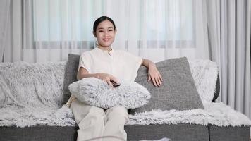 An optimistic girl smiling positively sitting in the house watching TV holding a remote control on the sofa. video