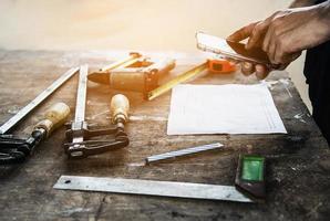 Carpenter working with wood furniture product preparation using  hand tools and machine table station while using mobile phone photo