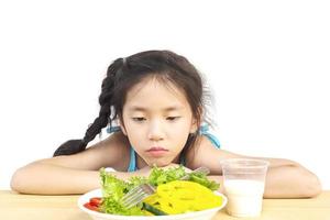 encantadora chica asiática que muestra una expresión aburrida con verduras frescas y coloridas y un vaso de leche aislado sobre fondo blanco foto