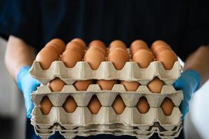 A man carry 3 layers eggs tray for stay safe at home putting sanitary glove during corona virus COVID-19 spreading - people keeping food in COVID-19 viral spread period concept. photo