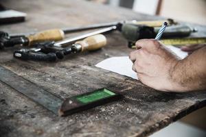 carpintero que trabaja con la preparación de productos de muebles de madera usando herramientas manuales y estación de mesa de máquina mientras escribe papel foto