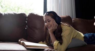 bella joven asiática usando una laptop y usando audífonos escuchando música y acostada en un sofá en la sala de estar foto