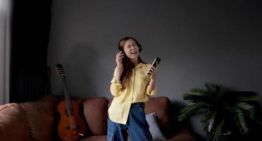 Young asian woman listening to music on couch in living room at home. Happy asia female using mobile smartphone, wearing headset photo