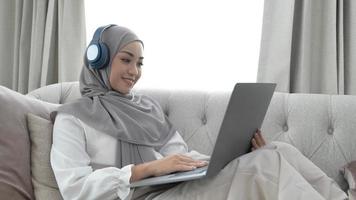 Charming young Asian Muslim woman wearing hijab sits on a comfy sofa, using a laptop computer and listening to music on her headphones. photo