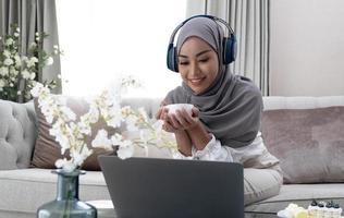 Domestic Lifestyle. Happy Muslim Woman Using Laptop At Home, Watching Movie And Drinking Coffee, Relaxing On Couch In Living Room, Side View photo