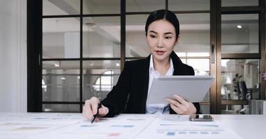 atractiva y encantadora joven empresaria asiática o trabajadora financiera en traje formal usando tableta y trabajando con su informe financiero en la oficina. foto