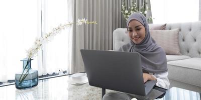 concepto de educación en línea mujer árabe sonriente con un pañuelo en la cabeza en su computadora portátil se sienta en la parte posterior del sofá en casa. disfrute del espacio de copia de aprendizaje a distancia foto
