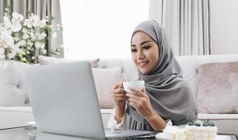 estilo de vida doméstico. mujer musulmana feliz usando una laptop en casa, viendo películas y tomando café, relajándose en el sofá en la sala de estar, vista lateral foto