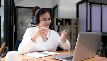 Smiling young asian woman in headset wave greet talking on webcam virtual conversation on laptop, happy female in wireless headphones speak on video call on computer, consult client online photo