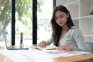 Portrait of asian business woman paying bills online with laptop in office. Beautiful girl with computer and chequebook, happy paying bills. Startup business financial calculate account concept photo